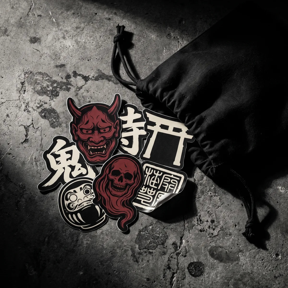 A die-cut IKITAI sticker pack — red oni mask, kanji and skull designs — beside a black drawstring cotton dust bag
